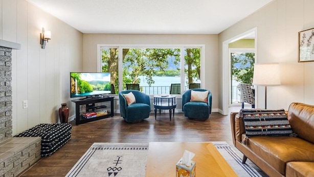 Ein Wohnzimmer mit Fernseher, zwei Sesseln, einer Couch und Seeblick durch große Fenster.