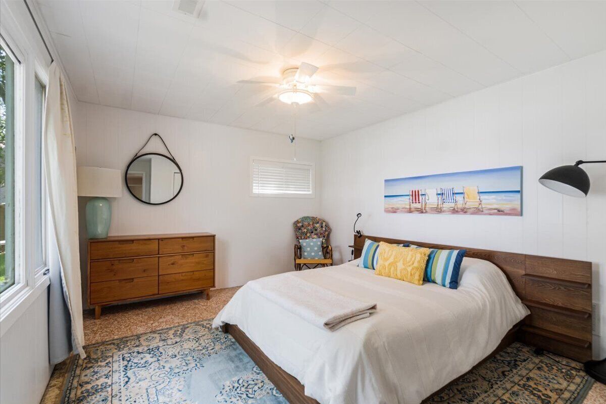 Un dormitorio con cama, cómoda, silla, ventilador de techo, ventana y un cuadro en la pared.