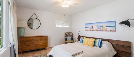 Un dormitorio con cama, cómoda, silla, ventilador de techo, ventana y un cuadro en la pared.
