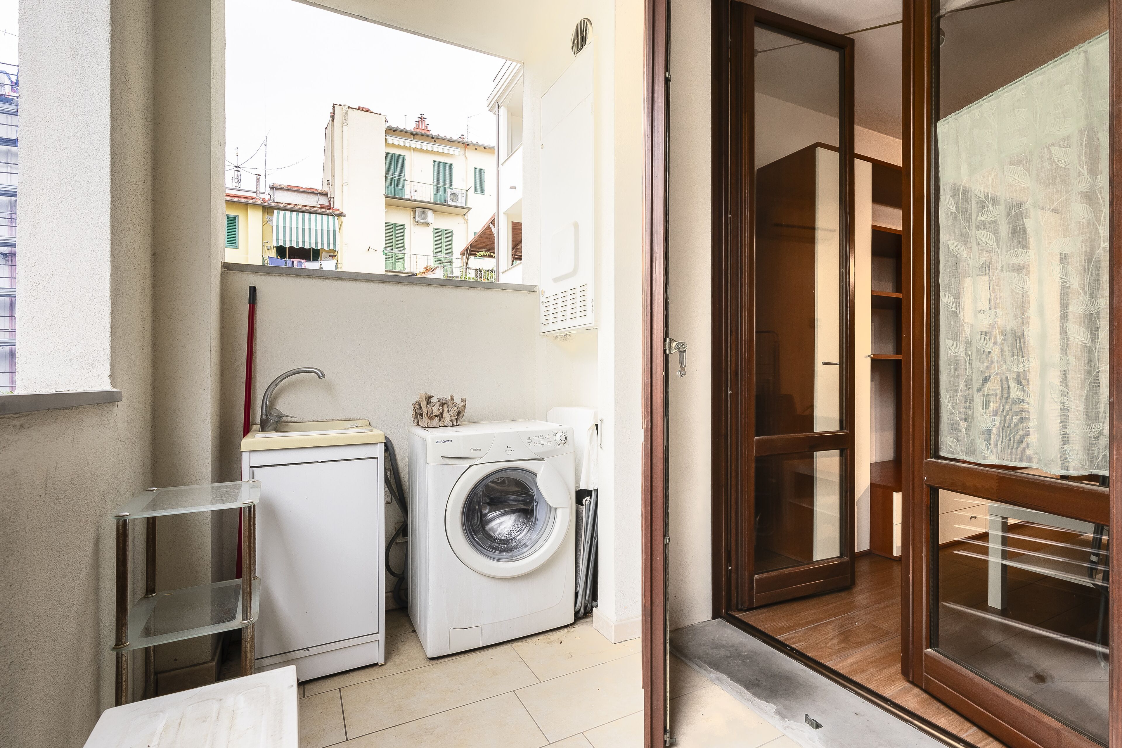 Un espace réduit dédié à la buanderie, comprenant un lave-linge, un étendoir, une petite étagère en métal et une vue sur les bâtiments par une porte ouverte.