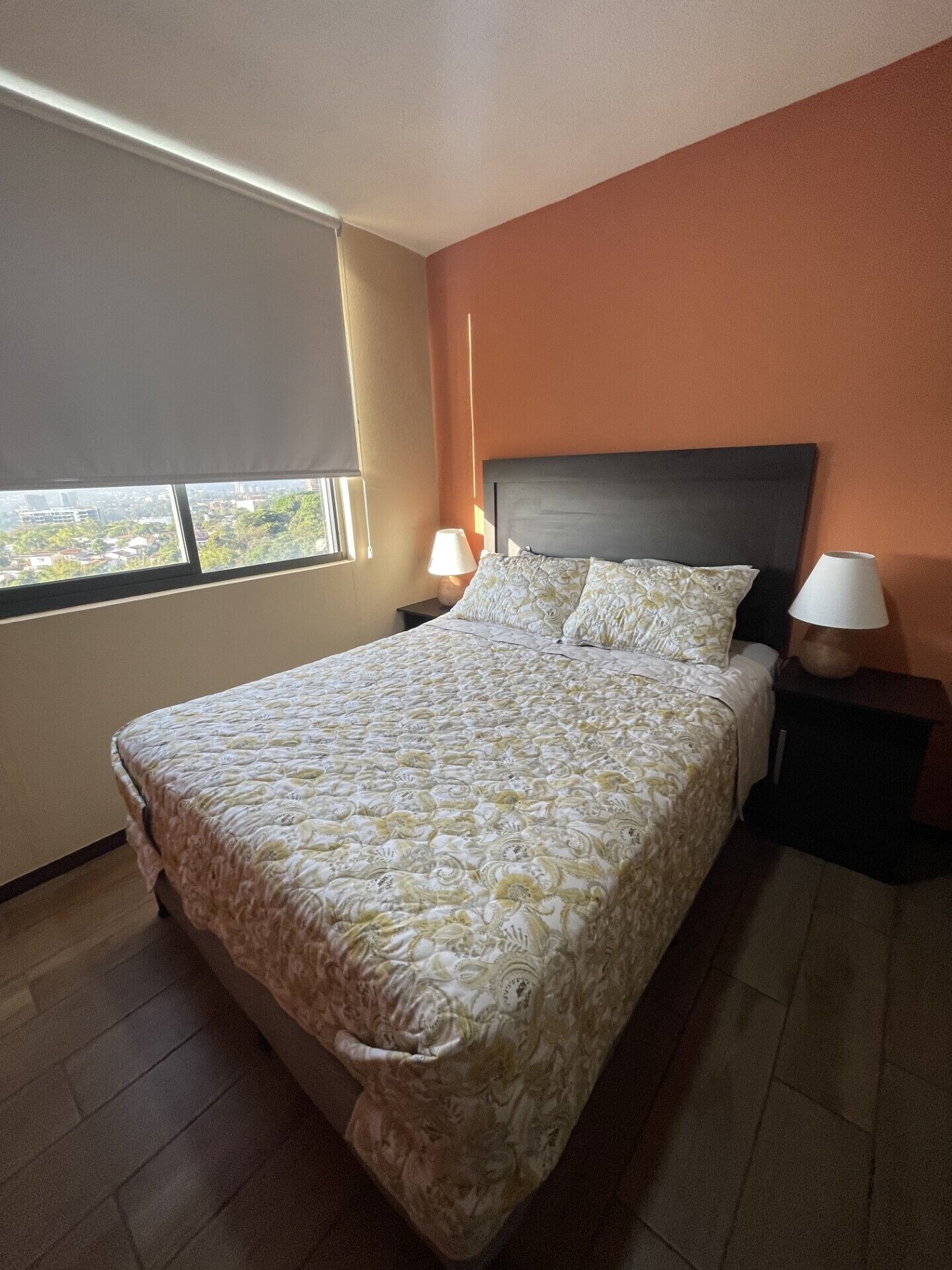 A bedroom with a bed, two bedside tables, a window, and a view of the city.