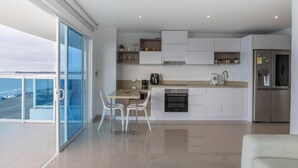 Eine moderne Küche mit einer großen Kochinsel, weißen Schränken und einem Edelstahlkühlschrank. Es gibt einen Essbereich mit Tisch und Stühlen, und ein großes Fenster bietet Meerblick.