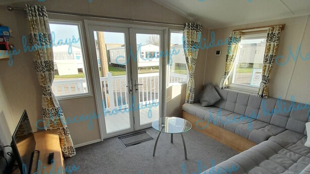 A cozy living room with a grey sofa, a small glass table, and a wooden side table. There are two large windows with curtains, and a view of the outside. - Pet Friendly Caravan in Walton on the Naze (Walton on the Naze)