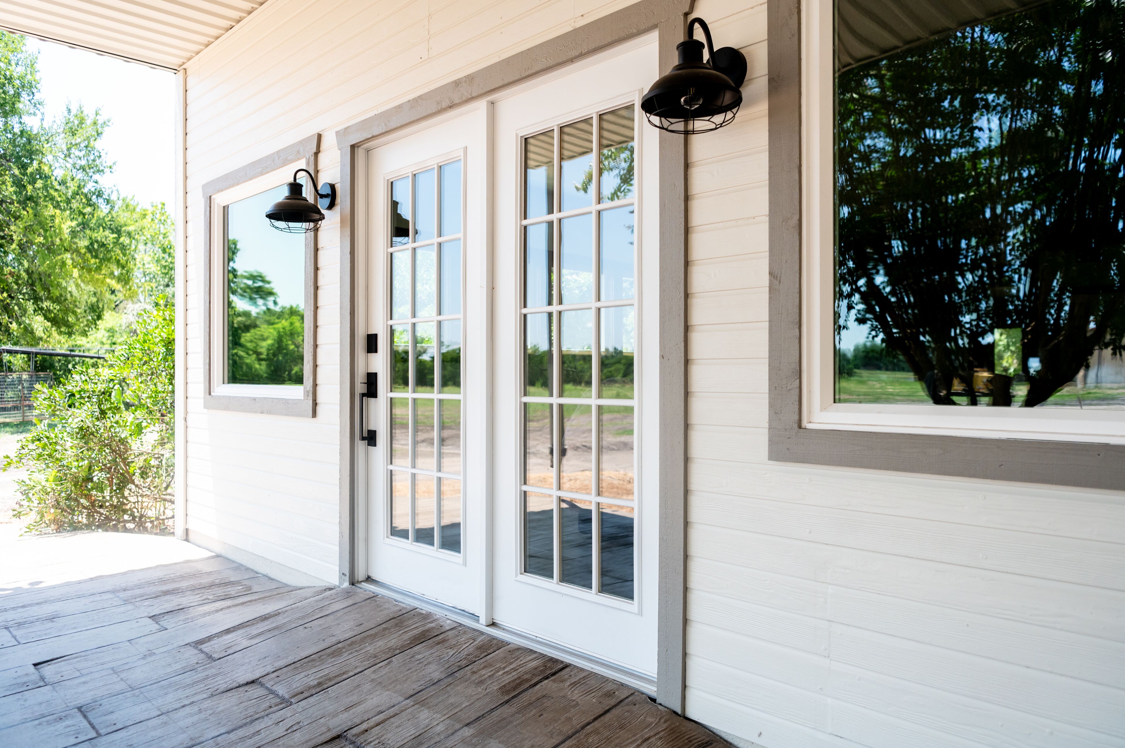 A covered porch with wooden flooring, white exterior walls, and large windows with a view of greenery.