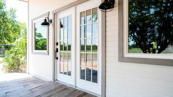 A covered porch with wooden flooring, white exterior walls, and large windows with a view of greenery.
