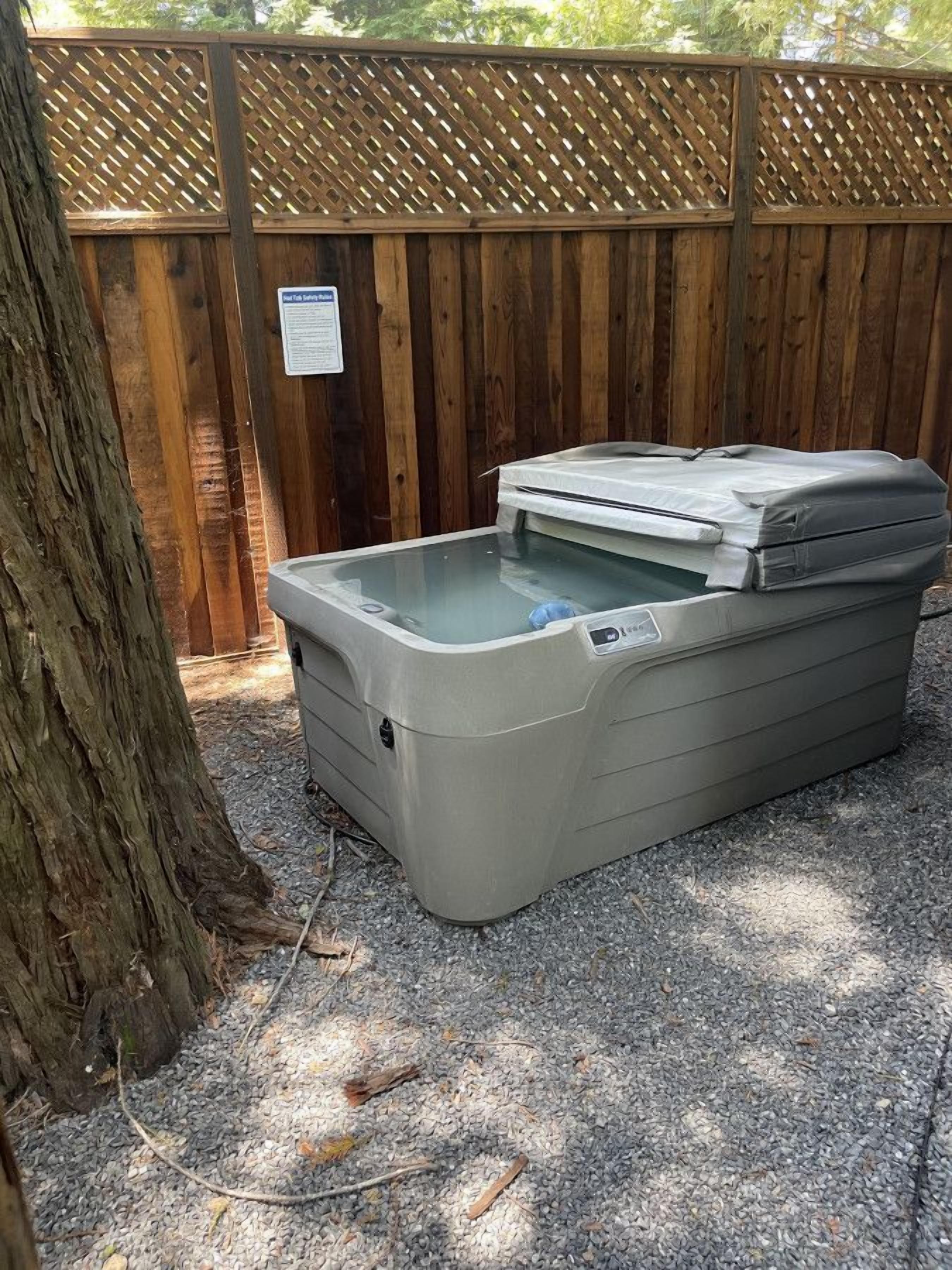 Un jacuzzi portátil con tapa, sobre suelo de grava, junto a una cerca de madera.