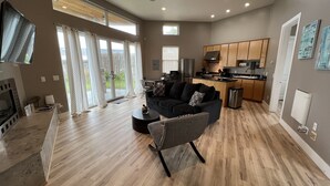 A spacious living area with a dark sofa, a small round coffee table, and a grey armchair. There is a kitchen area with wooden cabinets and a stainless steel refrigerator. Large windows with curtains provide natural light and a view of the outdoors.