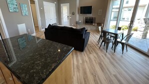 A modern living room with a black sofa, a dining area with a table and chairs, a flat-screen TV mounted on the wall, and a large sliding glass door leading to an outdoor patio.