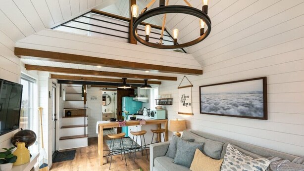 A spacious living area with a grey sofa, wooden beams, a kitchen in the background, and a view of the sea through a window.