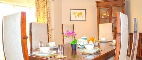 Une salle à manger avec une table en bois dressée pour quatre personnes, entourée de chaises rembourrées. Un lustre est suspendu au plafond et une carte du monde est accrochée au mur.