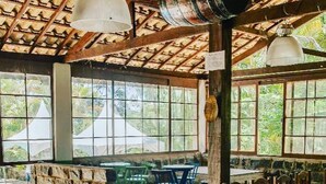 A dining area with a long table covered in red, surrounded by chairs, under a wooden ceiling with hanging lights.
