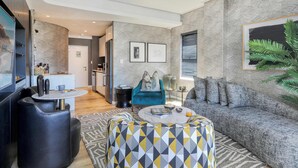 Ein modernes Wohnzimmer mit einer Couch, einem gestreiften Hocker und einem Couchtisch. Im Hintergrund ist eine Küche mit einem Kühlschrank und einer Arbeitsplatte zu sehen.