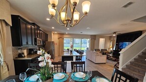 A dining area with a dark wooden table set for six, surrounded by chairs. A chandelier hangs above, and a kitchen is visible in the background. - Solara Resort private POOL HOME 4 Bed (Kissimmee)