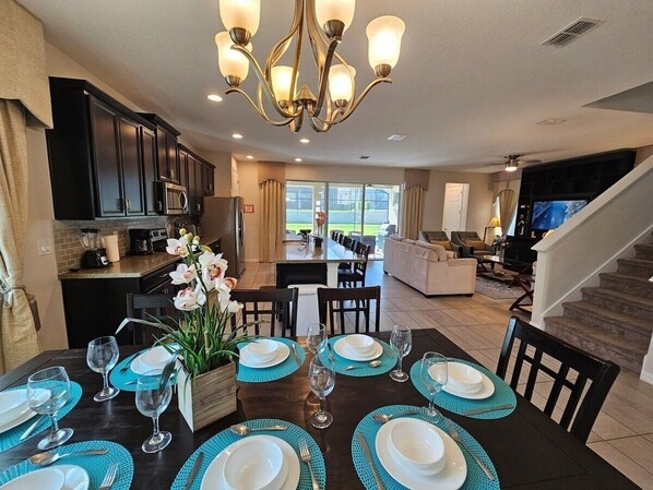 A dining area with a dark wooden table set for six, surrounded by chairs. A chandelier hangs above, and a kitchen is visible in the background.