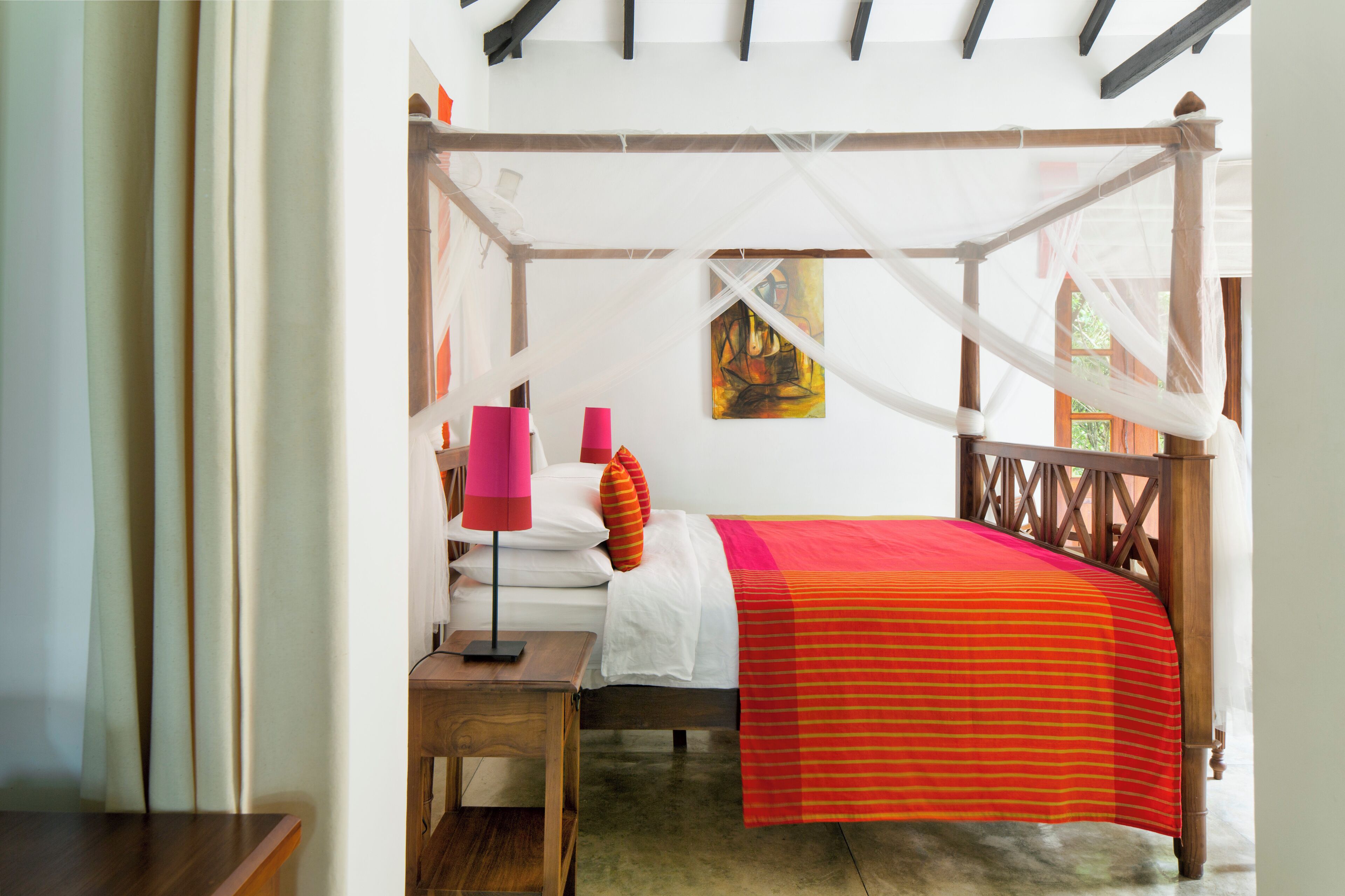 A four-poster bed with a canopy, a bedside table with a lamp, and a painting on the wall.