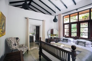 A bedroom with a large bed, a wooden headboard, a chair, a dresser, and a window with a view of greenery. - Rosyth Estate House (Pinnawala)