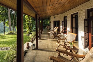 A covered outdoor seating area with wicker chairs and a table, overlooking a lush garden and a stone building with windows. - Rosyth Estate House (Pinnawala)
