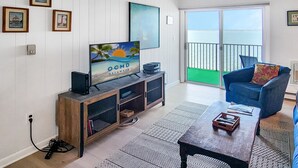 Een woonkamer met een televisie, een blauwe fauteuil, een kamerkleed met dessin en uitzicht op zee door een schuifpui.