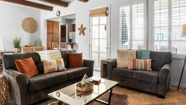 Ein Wohnzimmer mit zwei dunkelgrauen Sofas, einem Glastisch und einem großen Fenster mit Jalousien.