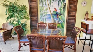 Zona pranzo con tavolo in vetro circondato da sedie in vimini con cuscini blu, una pianta in vaso di grandi dimensioni e un murale raffigurante una scena tropicale.
