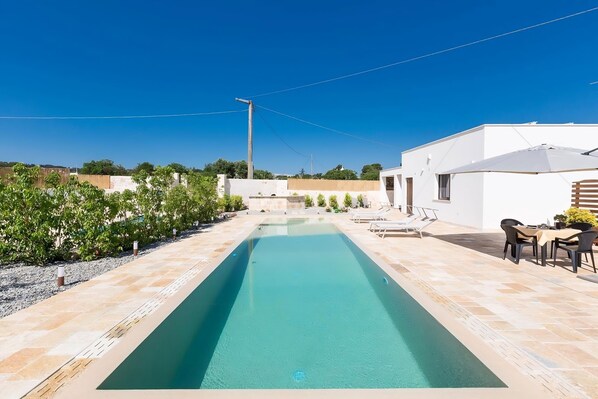 A rectangular swimming pool with a tiled deck, surrounded by greenery and a white building with a patio area.