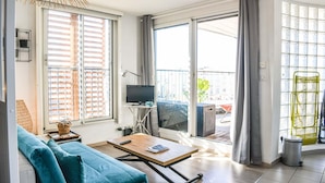 Un salon moderne avec un canapé, une table basse, un téléviseur et de grandes fenêtres équipées de stores.