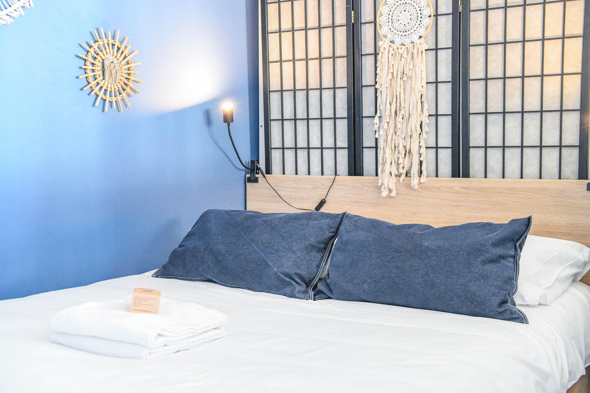 Ein ordentlich bezogenes Bett mit blau-weißer Bettwäsche, eine Nachttischlampe und eine Wand mit einem Dekorationselement sowie ein Fenster mit Gittermuster.