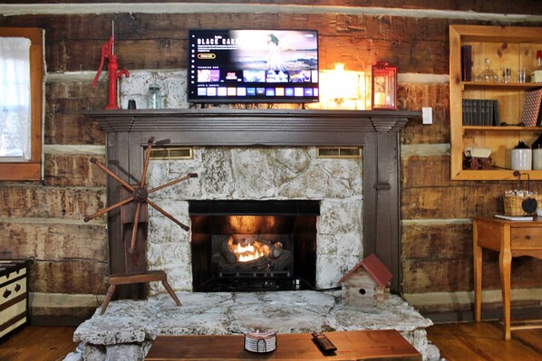A fireplace with a television above it, a wooden shelf with books and glassware, and a rustic stone wall. - Rustic Retreat, a pre Civil War log cabin. (Lucas)