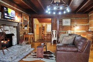 A cozy log cabin interior with a stone fireplace, leather sofa, wooden dining table, and a television mounted on the wall. - Rustic Retreat, a pre Civil War log cabin. (Lucas)