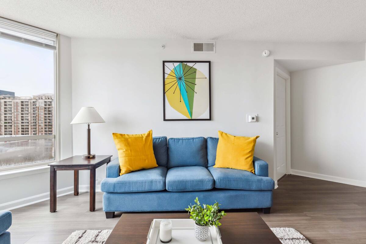 A modern living room with a blue sofa, yellow cushions, a framed abstract artwork, a small table with a lamp, and a plant on a tray.