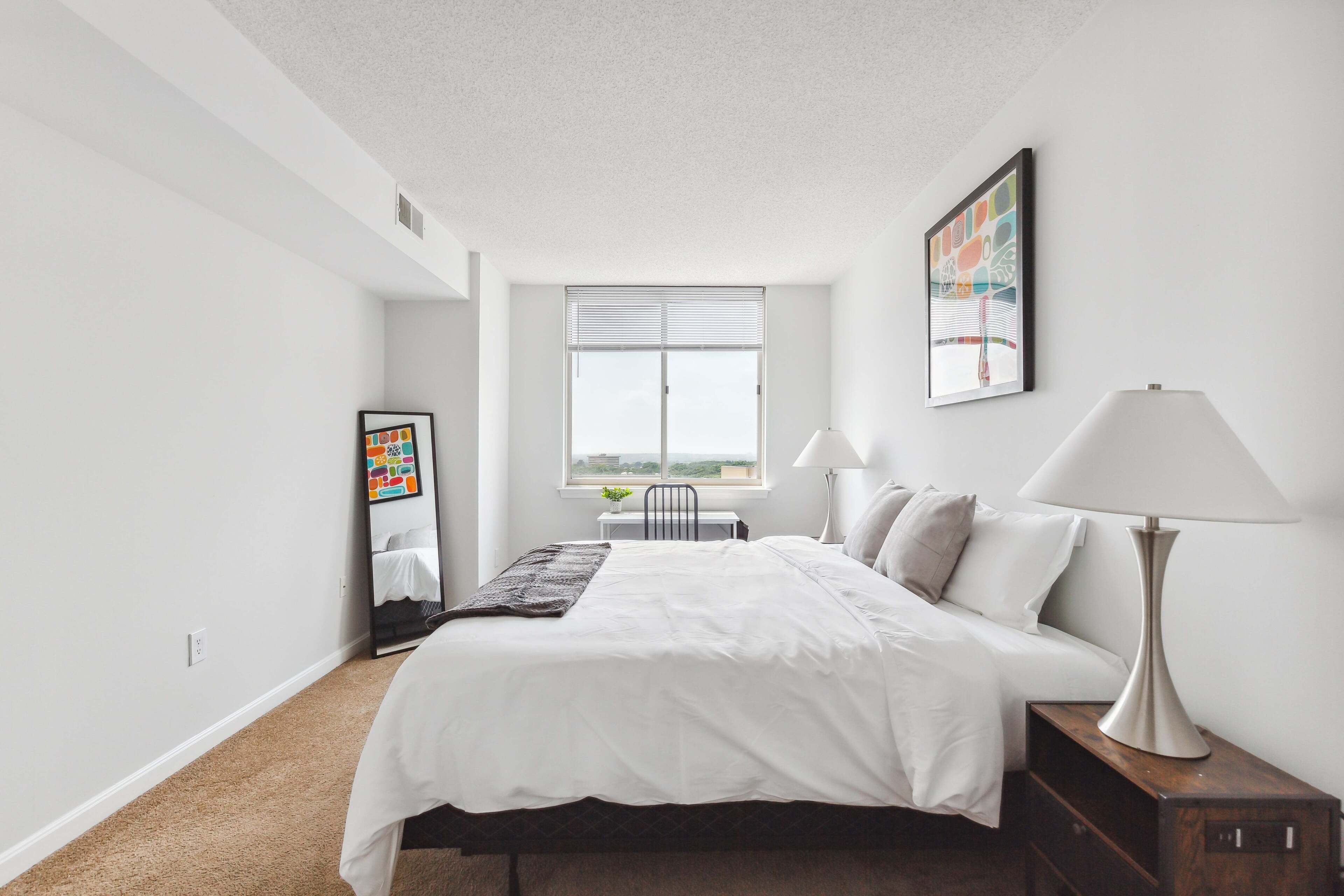 Un dormitorio moderno con una cama grande, una mesita de noche con lámpara, una ventana con persianas y un cuadro enmarcado en la pared.