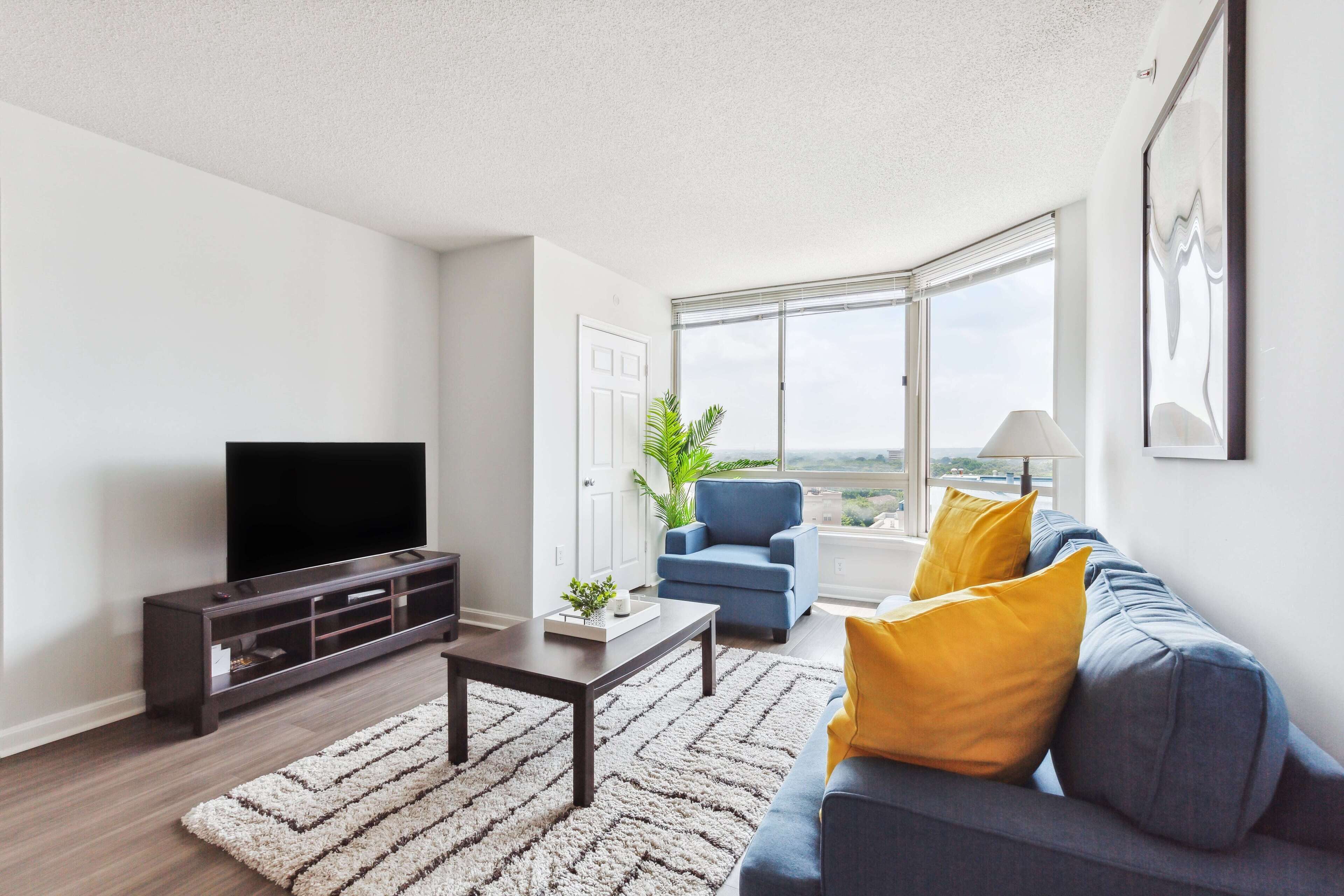 A modern living room with a flat-screen TV, a dark wooden entertainment unit, a blue armchair, a gray sofa with yellow cushions, and a large window offering a city view.