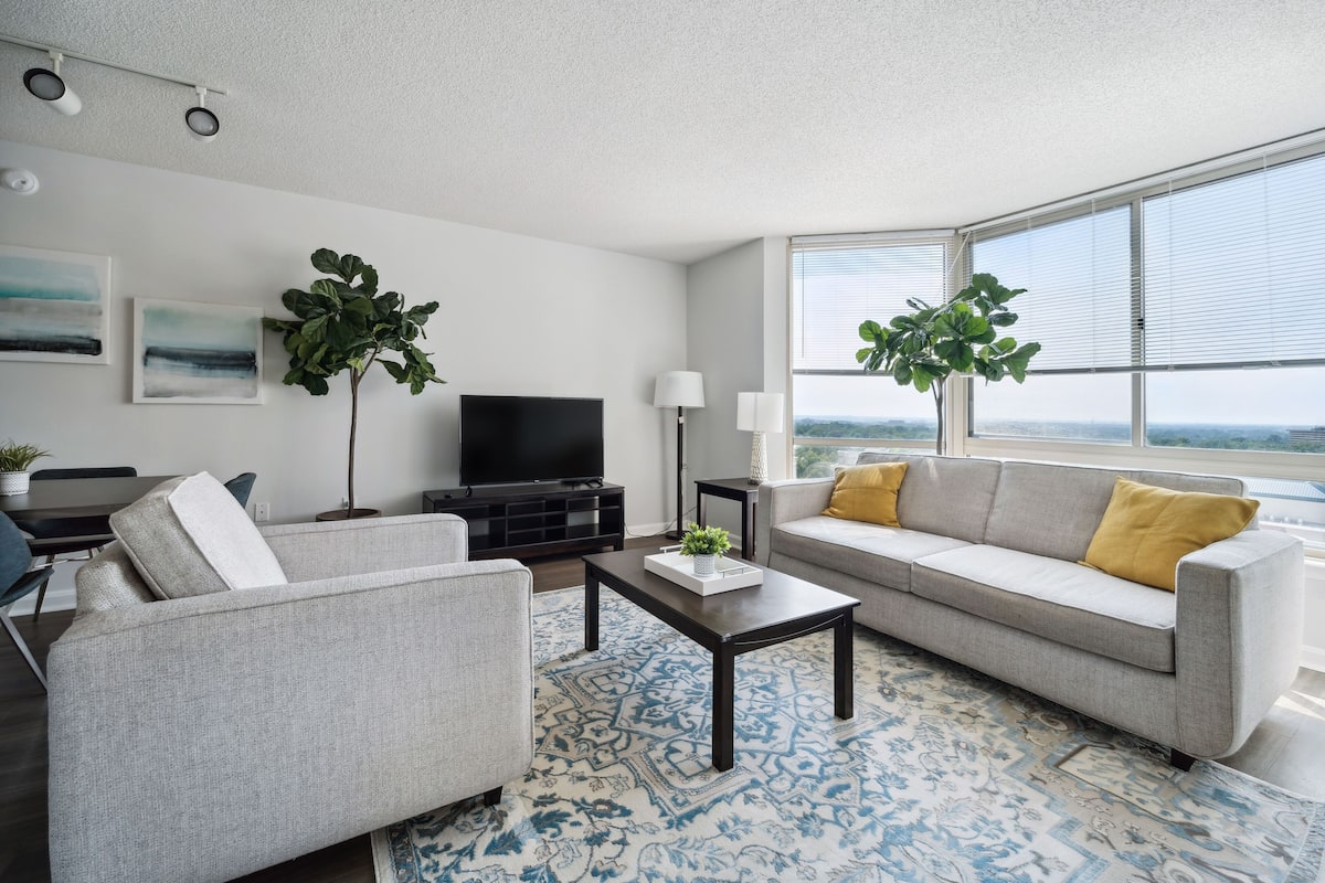 A modern living room with a grey sofa, a matching armchair, a coffee table, and a TV. There are two large windows with blinds, a plant, and artwork on the walls.