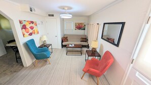 Sala de estar con sofá, dos sillones, una mesa de centro y una lámpara. Hay una puerta y un espejo en la pared.