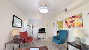 Habitación de hotel con televisor sobre un mueble, una silla roja, una silla azul, una lámpara y un cuadro de flores en la pared.