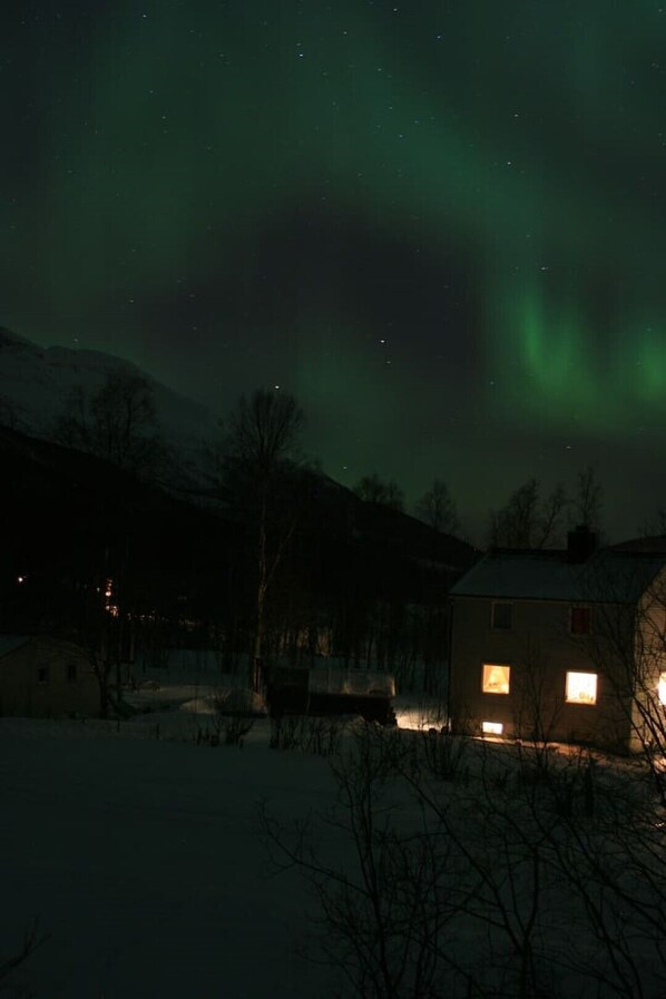 Una casa con una ventana iluminada, rodeada de nieve, bajo un cielo nocturno con estrellas visibles y auroras boreales.