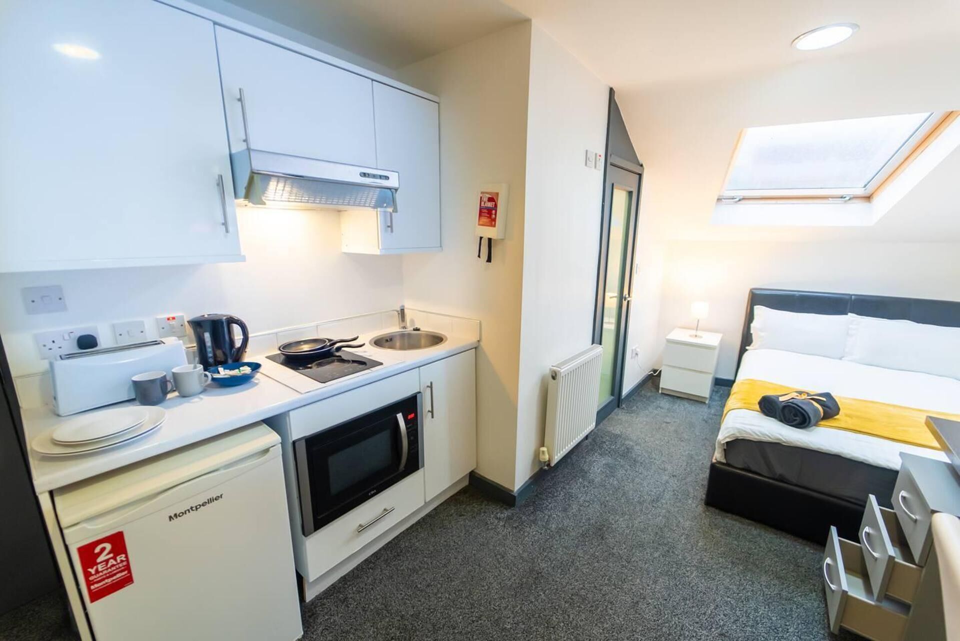 A compact hotel room with a kitchenette, a bed with a headboard, a small bedside table, and a window with a view.