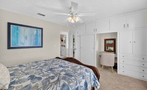 Une chambre à coucher avec un lit, un ventilateur de plafond, des armoires blanches et un tableau encadré au mur.
