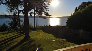 Un patio trasero con una terraza de madera, un césped y vista a un lago con árboles y un atardecer.