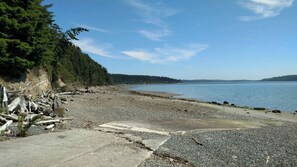 Una playa de piedras con un camino de hormigón que conduce al agua, rodeada de un bosque denso.