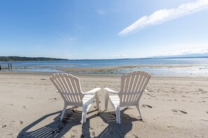 Two white Adirondack chairs facing a beach with a clear sky and distant mountains.