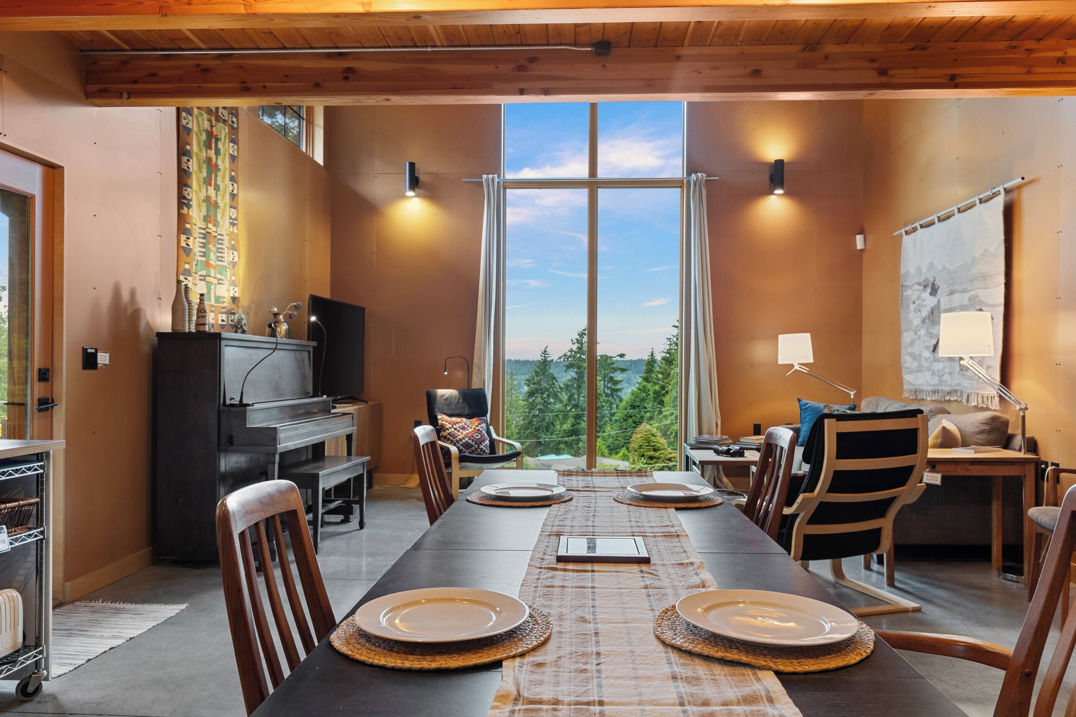 A dining area with a long table set for four, a piano, and a large window with a view of trees.