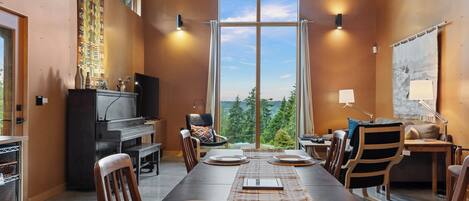 A dining area with a long table set for four, a piano, and a large window with a view of trees.