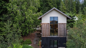 A modern cabin with a large glass window, surrounded by dense forest. - Crows Nest by Avantstay Accessible Home w/ Views (Clinton)