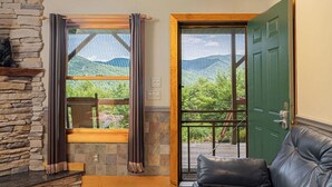 A room with a stone fireplace, a window with curtains, a green door, and a leather sofa. - Among the Clouds by Avantstay Views + Hot Tub (Candler)