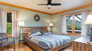 A bedroom with a wooden bed, two bedside tables with lamps, a chair, a ceiling fan, and a view of greenery through the windows. - Among the Clouds by Avantstay Views + Hot Tub (Candler)