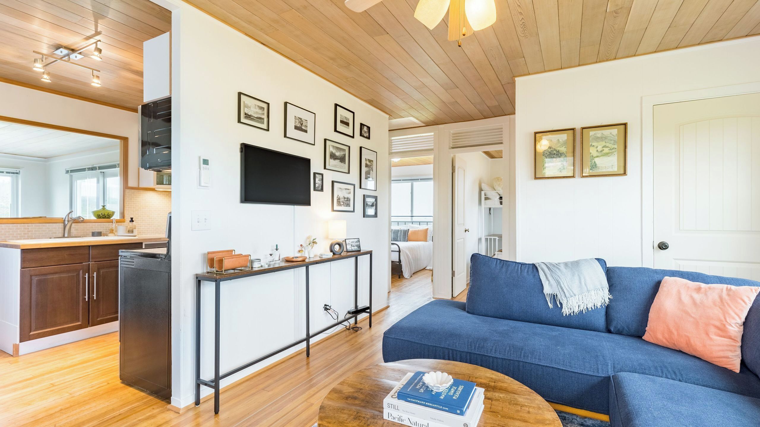 A modern living room with a blue sofa, a coffee table with a book, a kitchen with wooden cabinets, and a dining area with a white table and chairs.