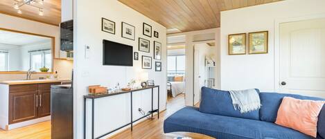 A modern living room with a blue sofa, a coffee table with a book, a kitchen with wooden cabinets, and a dining area with a white table and chairs.