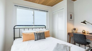 A bedroom with a bed, a desk, a chair, a window with a view, and a ceiling with wooden beams. - Swan Lake Cottage by Avantstay Views + Firepit (Oak Harbor)