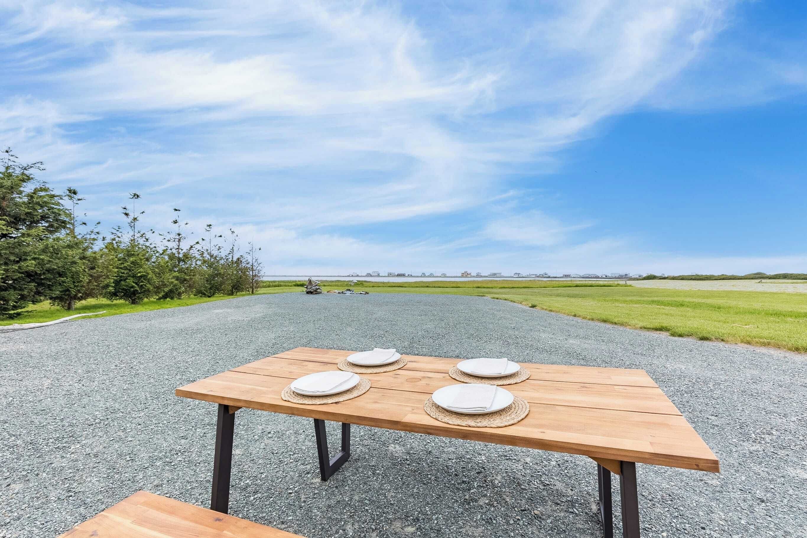 A wooden picnic table with four plates and a bench, set on a gravel path with a grassy field and trees in the background.